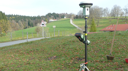GNSS-Empfänger auf einem Lotstab vor dem Hintergrund einer bayerischen Voralpenlandschaft mit Wiesen, Bergen und Bäumen.
